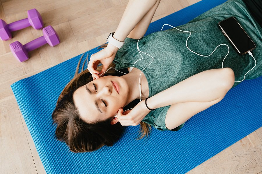 Ways to Modify Focus T25 Workouts Woman Putting Earbuds in Her Ears Laying Down on a Yoga Mat Next to a Small Pink Dumbbell