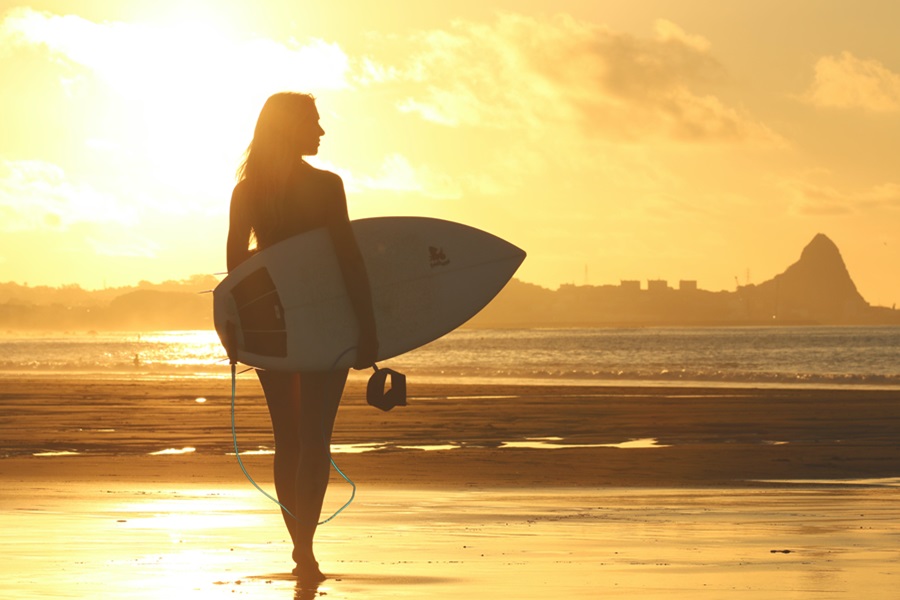 Things To Do in Los Angeles for Families a Woman Standing on a beach Holding a Surfboard with the Sun Setting in the Background