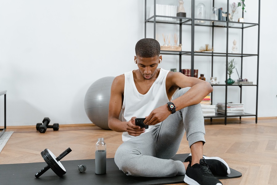 Jillian Michaels BODYSHRED Rise Workout Tips a Man Wearing Workout Clothes Sitting on the Floor Looking at His Phone with Dumbbells and a Water Bottle Next to Him
