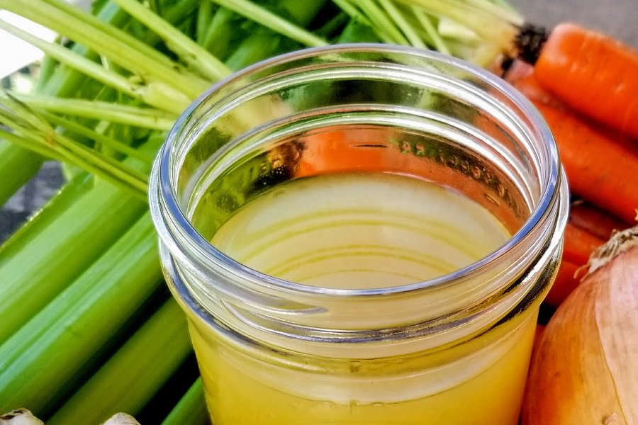 Crock Pot Bone Broth Recipe Close Up of a Jar of Bone Broth Surrounded By Veggies Including Celery, Carrots, and Ginger