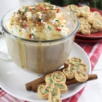 Gingerbread Latte Recipe Close Up of an Overflowing Cup of Latte on a Small Saucer with Two Gingerbread Cookies and Two Cinnamon Sticks