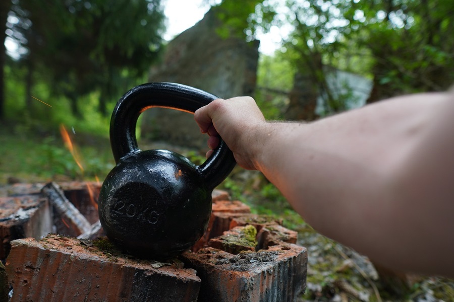 Kettlebell Exercises You Should Do Each Day Close Up of a Person's Hand Reaching for a Kettlebell on a Tree Stump in a Woodsy Area