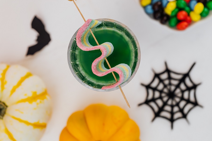 Halloween Margarita Drinks & Halloween Cocktail Recipes Overhead View of a Cocktail with a Sour Candy Ribbon Garnishing the Glass Next to a Small Pumpkin, a Plastic Spiderweb, and Halloween Candy