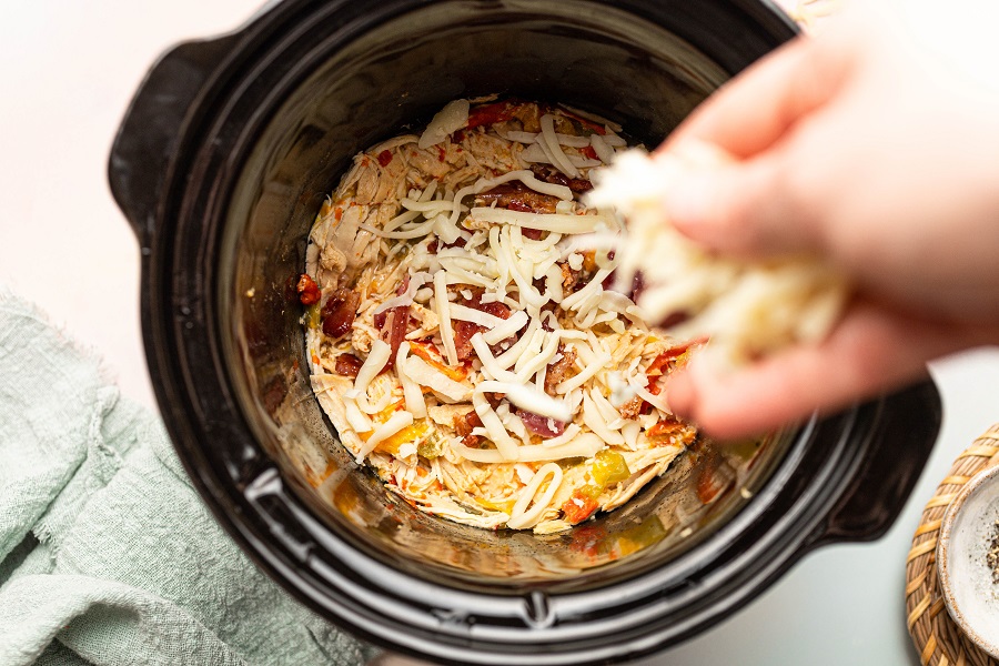 Crockpot Chicken and Bacon Keto Overhead View of a Crockpot with Chicken and Bacon Mixture Inside and a Person's Hand Sprinkling Shredded Cheese Into the Pot