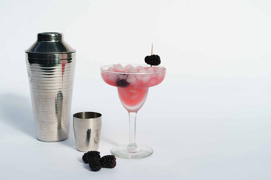 Margarita Pitcher Recipes that are Perfect for Parties a Single Pink Margarita Next to a Cocktail Shaker on a White Surface with a White Background