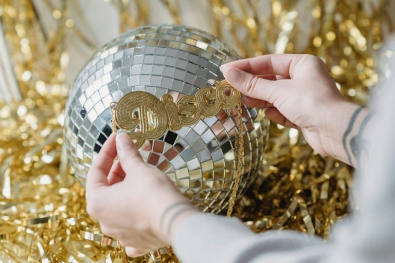 DIY New Years Decorations Close Up of a Person's Hands Holding a Golden Sign That Says Disco Over a Disco Ball with Some Gold Streamers in the Background