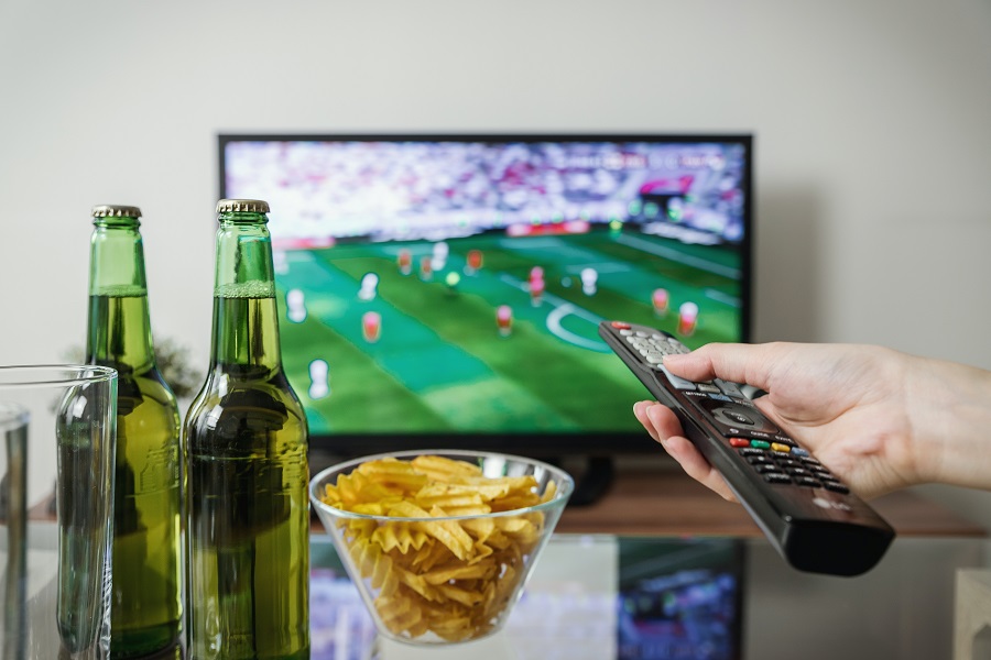 Ultimate Super Bowl Food Ideas List View of a Person's Hand Holding Out a TV Remote Pointing it at the TV with a Game Playing