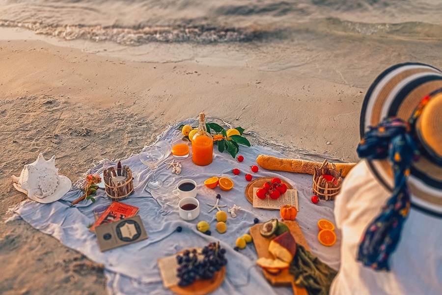 Beach Bag Essentials a Woman Sitting on a Beach Blanket Covered in Snacks Like Fruit, Cheese, and Bread