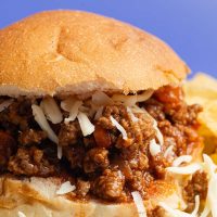 Easy Slow Cooker Sloppy Joes Recipe Close Up of a Sloppy Joe Sandwich with White Shredded Cheese and Blurred Potato Chips in the Background