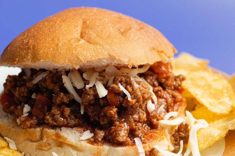 Easy Slow Cooker Sloppy Joes Recipe Close Up of a Sloppy Joe Sandwich with White Shredded Cheese and Blurred Potato Chips in the Background