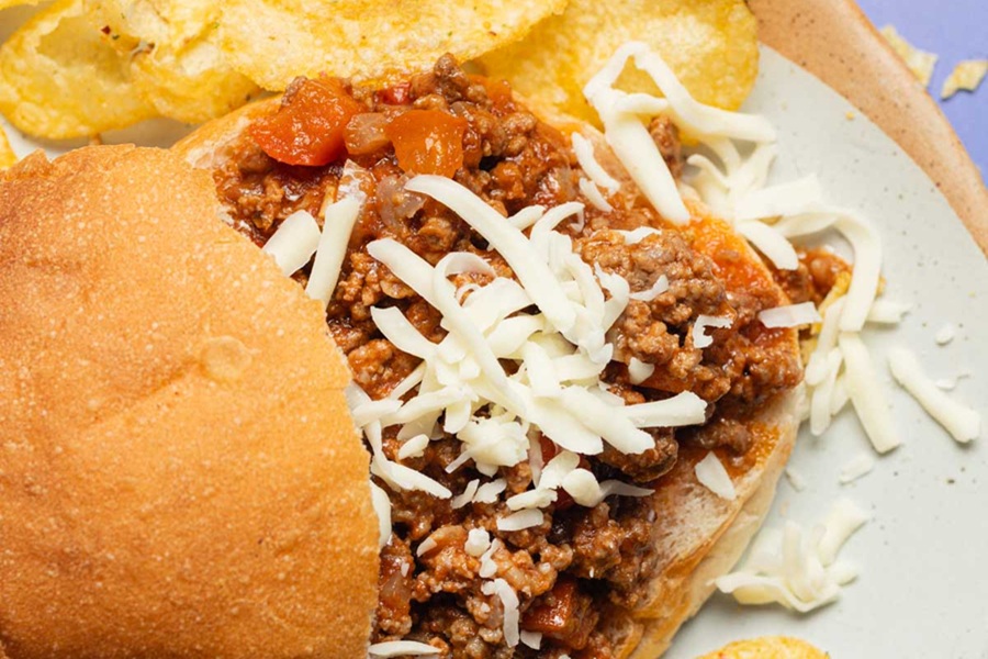 Easy Slow Cooker Sloppy Joes Recipe Close Up of a Sloppy Joe with the Top Bun Slightly Off Center White Shredded Cheese On Top of the Meat Mixture and Some Potato Chips on the Side