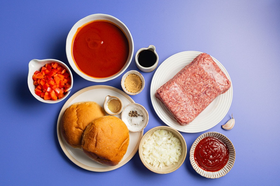Easy Slow Cooker Sloppy Joes Recipe Overhead View of Sloppy Joe Ingredients Divided Onto Plates Including Raw Meat, Burger Buns, Seasonings, Onions, Ketchup, Tomato Sauce, Diced Tomatoes, and Worcestershire Sauce