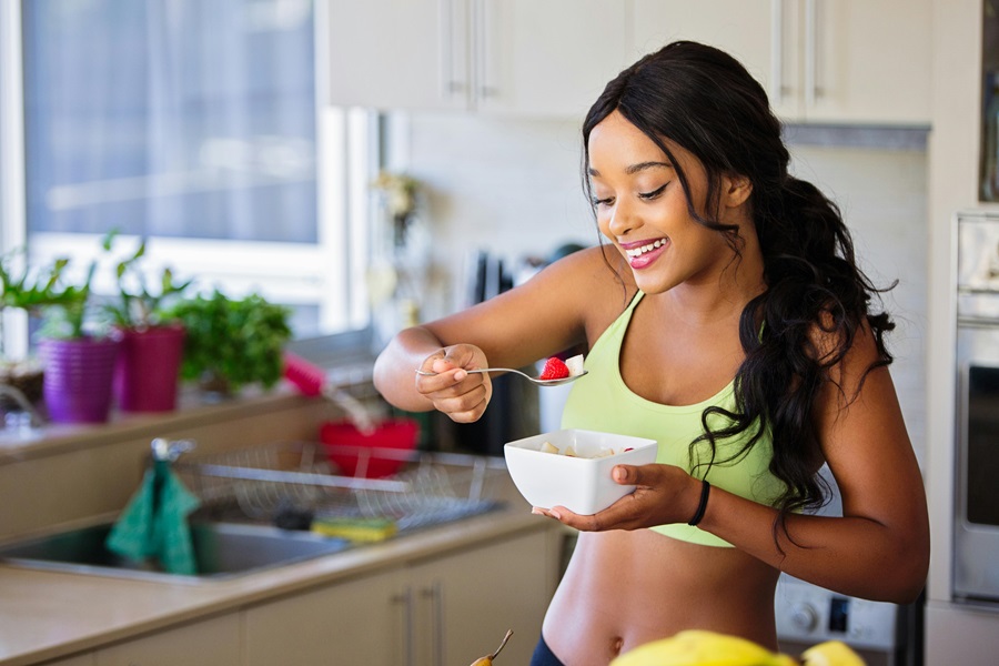 How to Get your Body in Ketosis a Woman Wearing Workout Clothes Standing in a Kitchen Eating a Bowl of Fruit and Yogurt