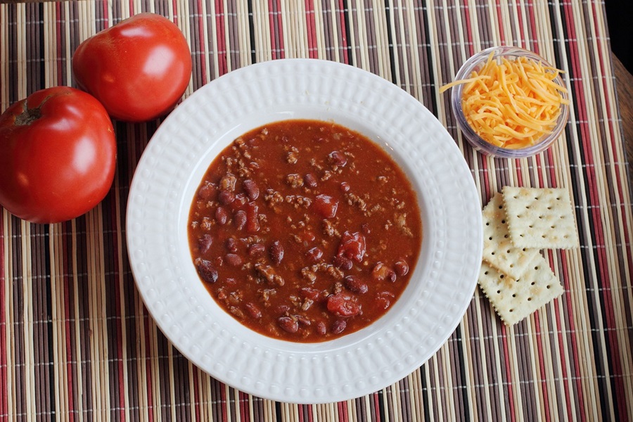 Keto Crockpot Chili Recipes Overhead of a Bowl of Chili Next to Tomatoes, Crackers, and Shredded Cheddar Cheese