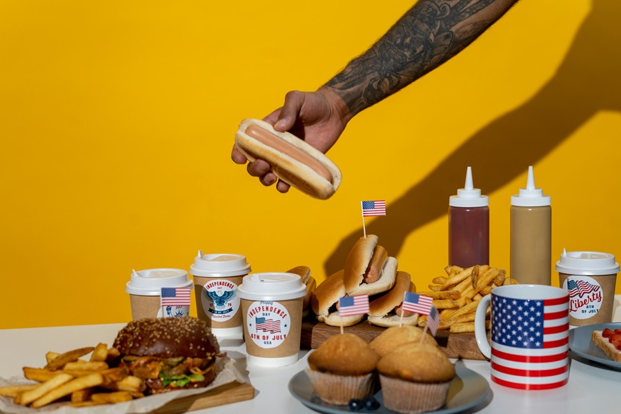 Easy 4th of July Recipes a Person's Hand Reaching Out Grabbing a Hot Dog From a Table of Fourth of July Food on a Table
