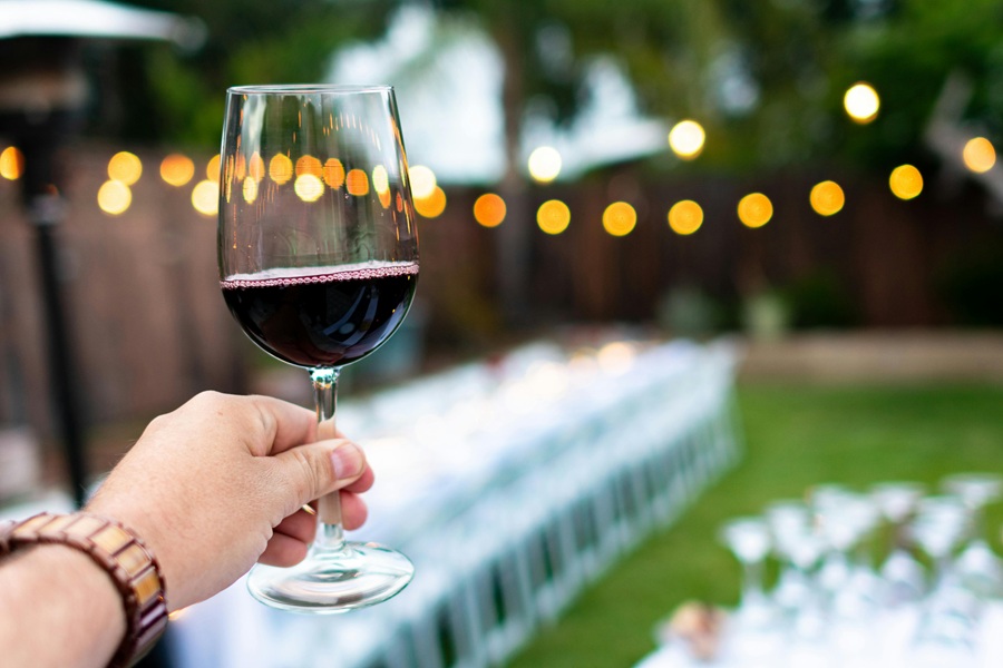 Fall Red Wine Sangria Recipes Close Up of a Person's Hand Holding a Glass of Red Wine Outside with a Blurred View of a Party Setting in the Background