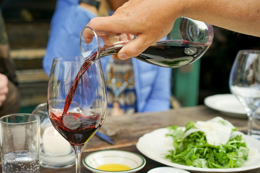 Fall Red Wine Sangria Recipes Close Up of a Person's Hand Pouring a Pitcher of Red Wine into a Wine Glass on a Wooden Table