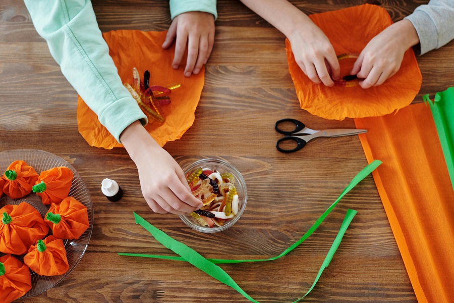 Crockpot Halloween Recipes Overhead View of Two Kids Making Halloween Candy Bundles with Orange Tissue Paper and Green Ribbon