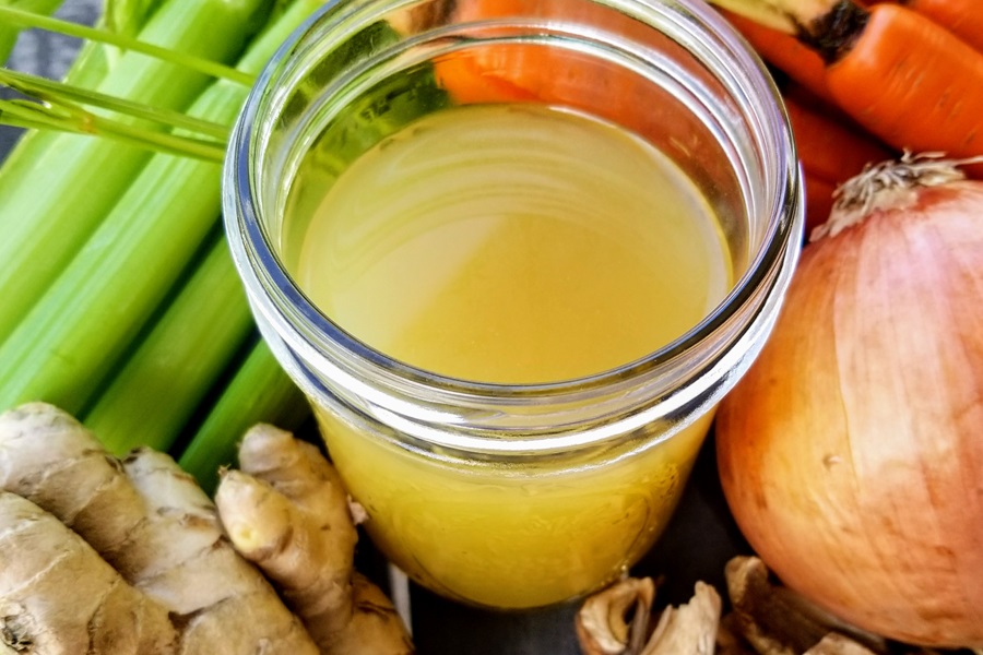 Healing Chicken Bone Broth Recipe Close Up of an Open Jar Filled with Broth Surrounded by Veggies