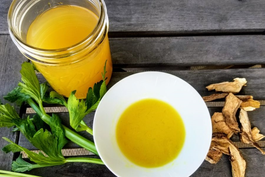 Healing Chicken Bone Broth Recipe Overhead View of a Bowl of Broth Next to a Glass Jar of broth on a Wooden Surface