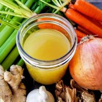 Healing Chicken Bone Broth Recipe Close Up of a Glass Jar Filled with Broth Surrounded by Carrots, Onions Ginger and Celery