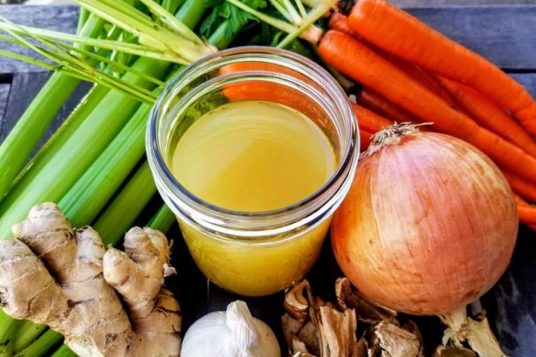 Healing Chicken Bone Broth Recipe Close Up of a Glass Jar Filled with Broth Surrounded by Carrots, Onions Ginger and Celery