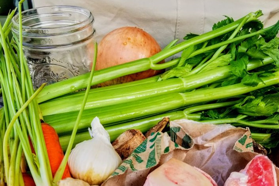 Healing Chicken Bone Broth Recipe Ingredients Laid Out on a Counter Top Including Celery, Carrots. Onions, Garlic, Chicken Bones, Mushrooms, and a Glass Jar