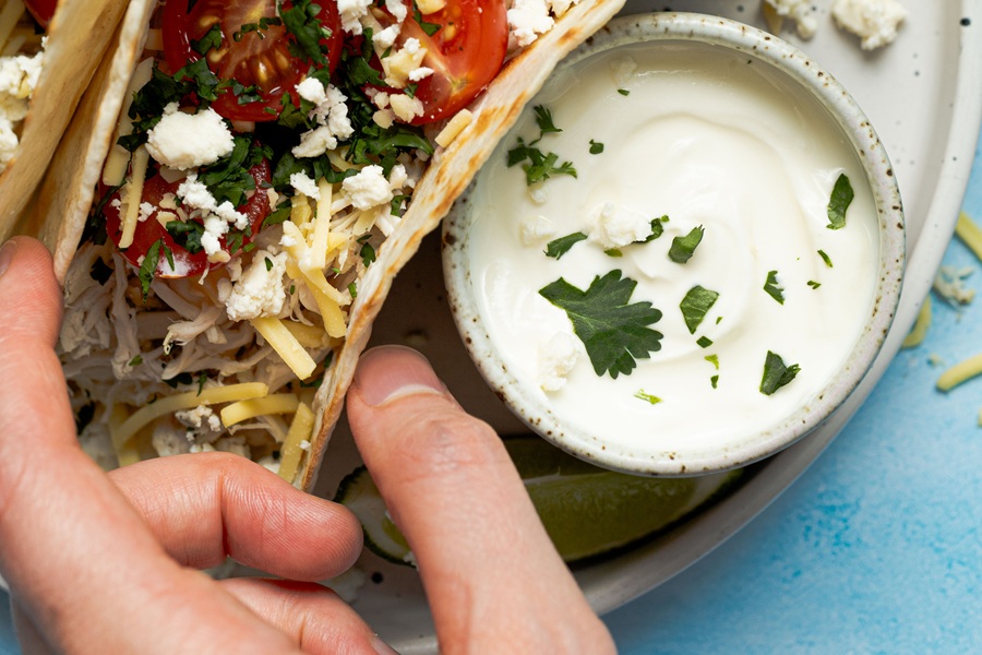 Easy Chicken Tacos with Shredded Chicken a Person's Hand Reaching for a Taco on a Plate with a Small Ramekin of Sour Cream Next to it