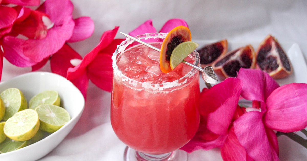 National Margarita Day Recipes Close Up of a Blood Orange Margarita Garnished with Dried Blood Orange Slices and a Flower Necklace Wrapped Around the Bottom of the Glass