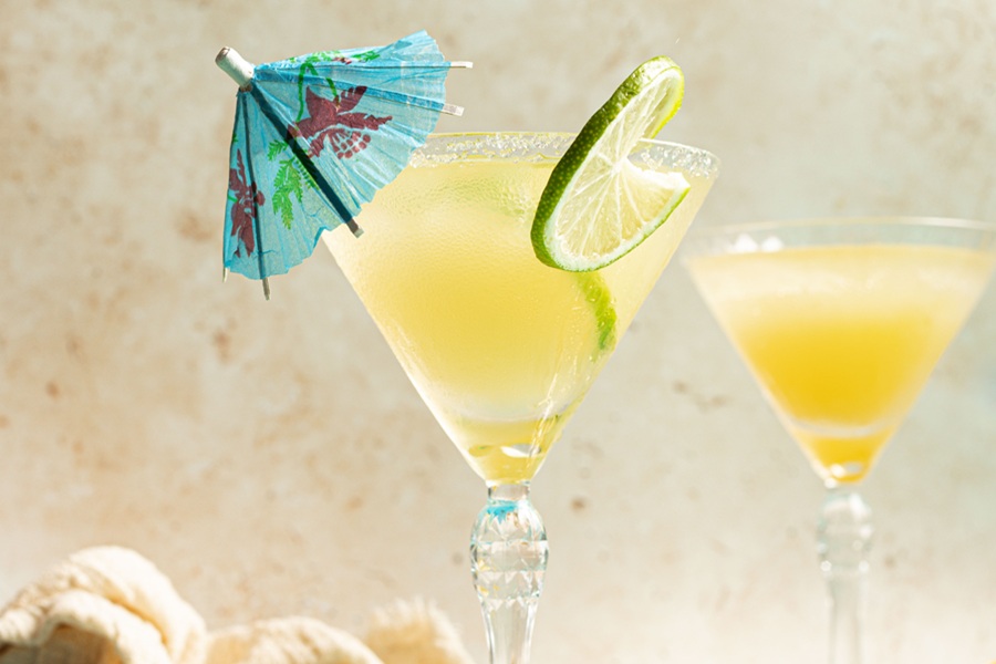 National Margarita Day Recipes a Margarita in a Martini Glass Garnished with a Lime Slice and a Small Blue Cocktail Umbrella with Another Cocktail in the Background