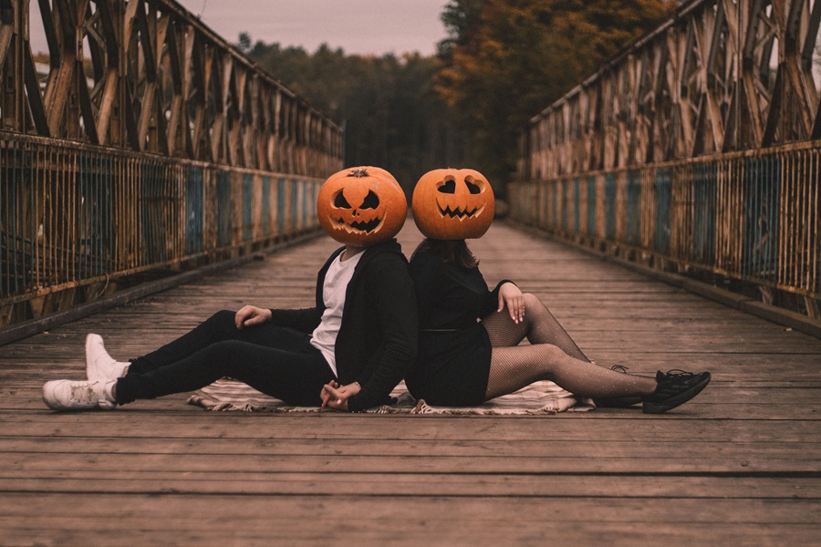 Downloads for your Halloween Playlist a Couple Sitting Back to Back on a Bridge in a Portrait Pose Each Wearing a Jack-O-Lantern on Their Heads