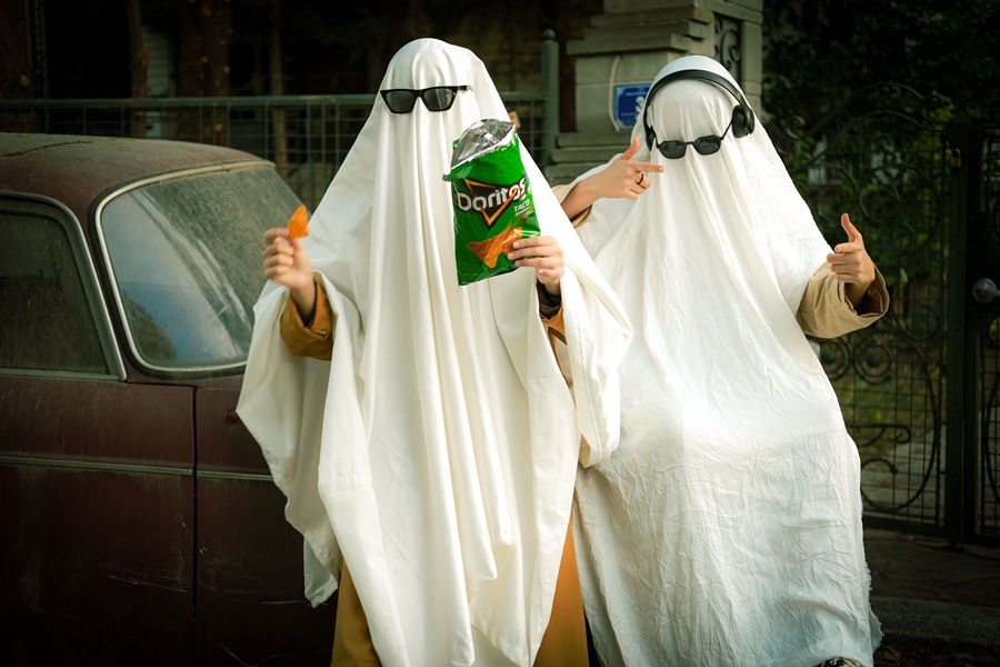 Downloads for your Halloween Playlist Two People Wearing White Sheets and Sunglasses One with Headphones on and the Other Holding a Bag of Chips