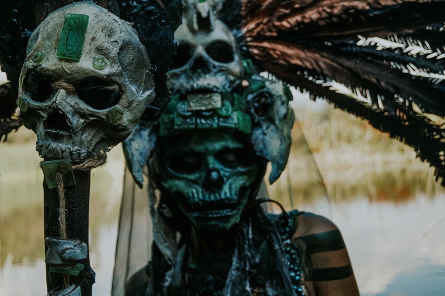 Downloads for your Halloween Playlist a Person Wearing Face Makeup to Look Like a Skeleton Holding a Skull Staff and Wearing a Headdress with a Skull at the Center and Feathers Splayed From it
